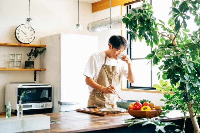 キッチンで料理をするエプロン姿の男性。妊娠中のパートナーをサポートするために食事を準備している様子。つわりで料理がつらい時に、家族やパートナーが食事作りを手伝うイメージ