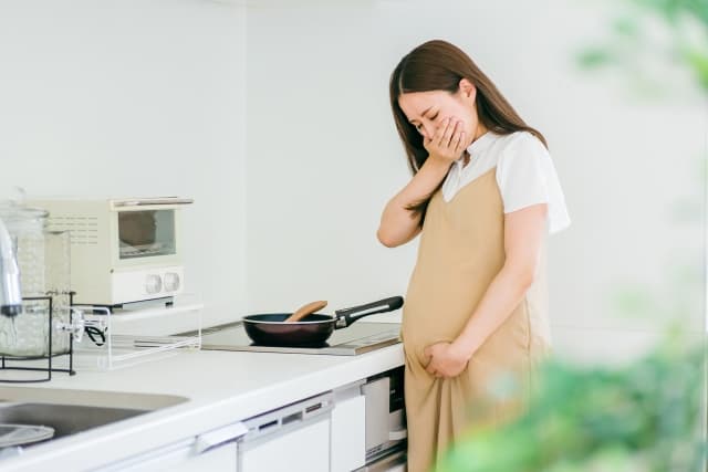 妊娠中の女性がキッチンで口元を押さえ、つわりによるにおいの不快感を感じている様子。料理中のにおいが辛くなる「においづわり」の例として、食事の準備が難しくなる状況を表している。